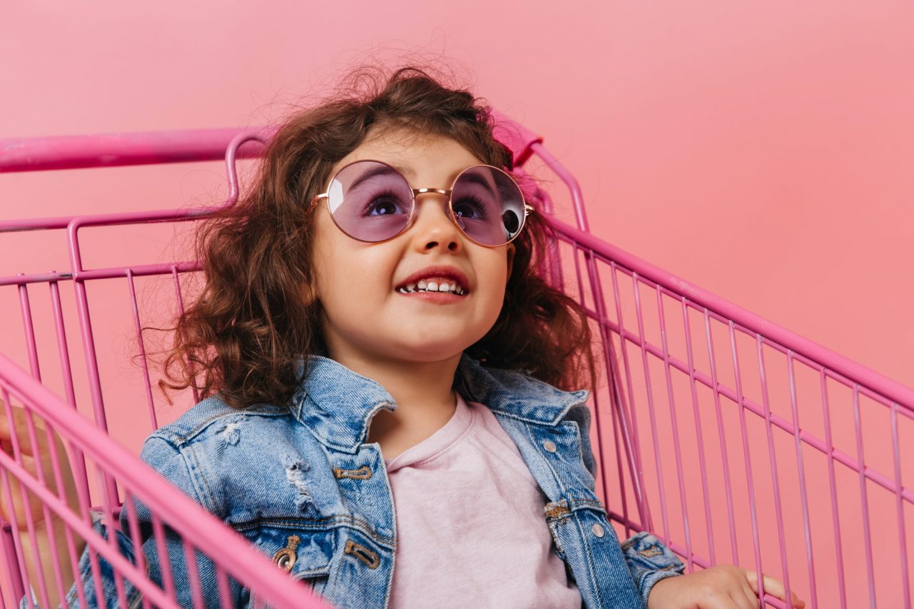preschooler girl sitting in shopping cart studio shot of laughing brunette kid in sunglasses .jpg